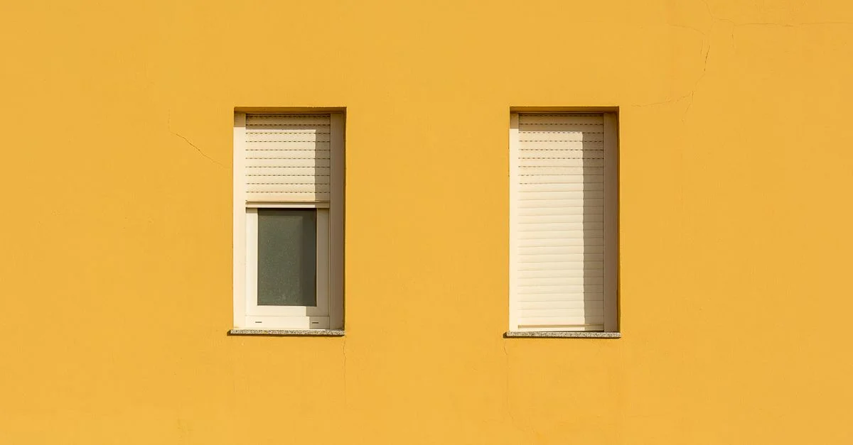 Un mur jaune avec deux fenêtres fermées avec des volets roulants, mettant en avant un design minimaliste.