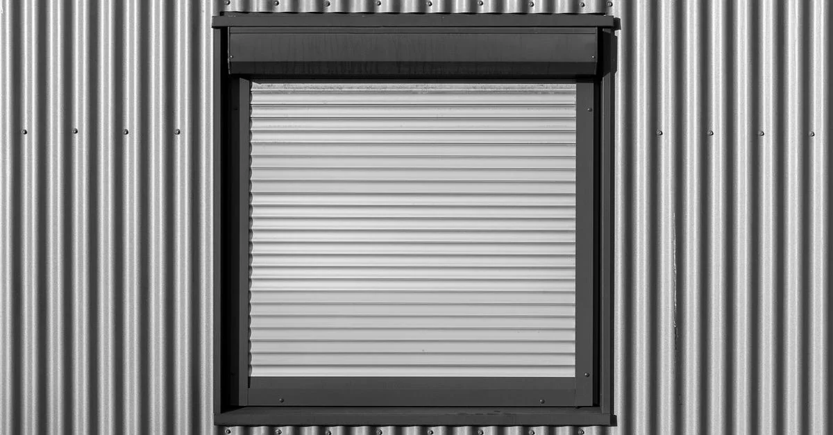 Photographie en noir et blanc d'une fenêtre avec volet roulant sur un mur en tôle ondulée, mettant en valeur l'architecture moderne.