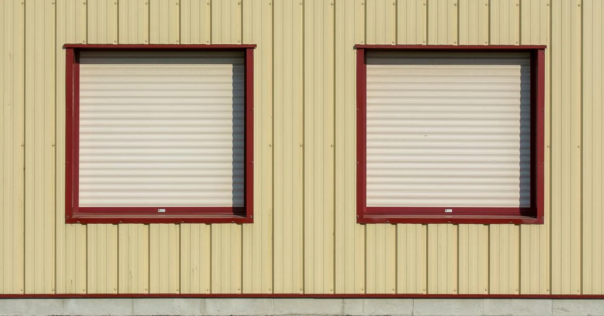 Deux volets roulants fermés sur la façade d'un bâtiment industriel jaune, avec des entourages rouges.
